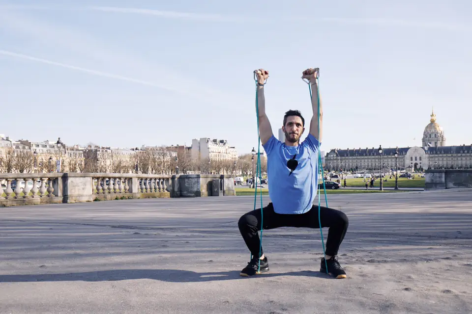 Coach sportif en squat avec élastiques, bras tendus, sur l'esplanade des Invalides